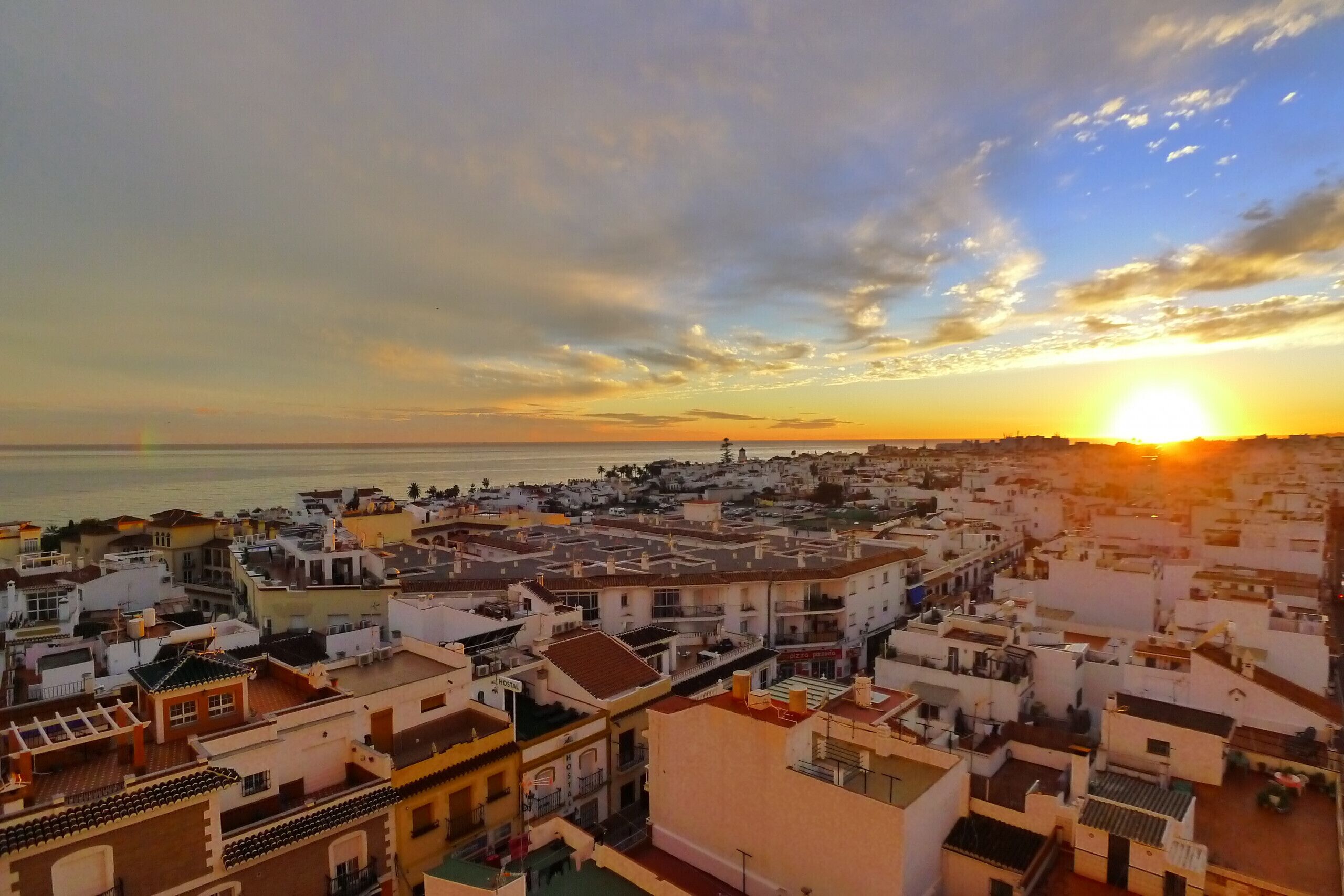 A coastal cityscape at sunset with buildings and a view of the sea.