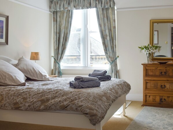 Un lit bien fait, recouvert d’une couvre-lit à motifs floraux, une commode en bois avec un miroir et une fenêtre agrémentée de rideaux.