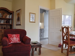 A living room with a red armchair, a wooden side table, a bookshelf, and a door leading to a kitchen.