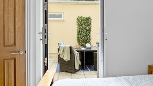 A room with a bed, a small table with a chair, and a view of an outdoor area with a wall-mounted plant and a window.