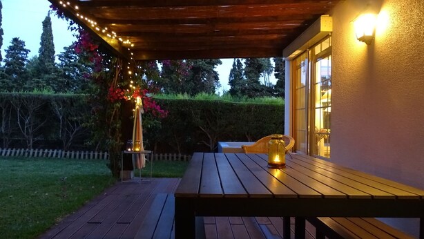 Een houten terras met een tafel en stoelen, sfeerverlichting en een decoratieve bloemstelling.