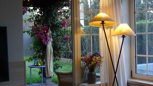 Een kamer met een groot raam, een rieten stoel, een klein tafeltje met een vaas en twee vloerlampen.