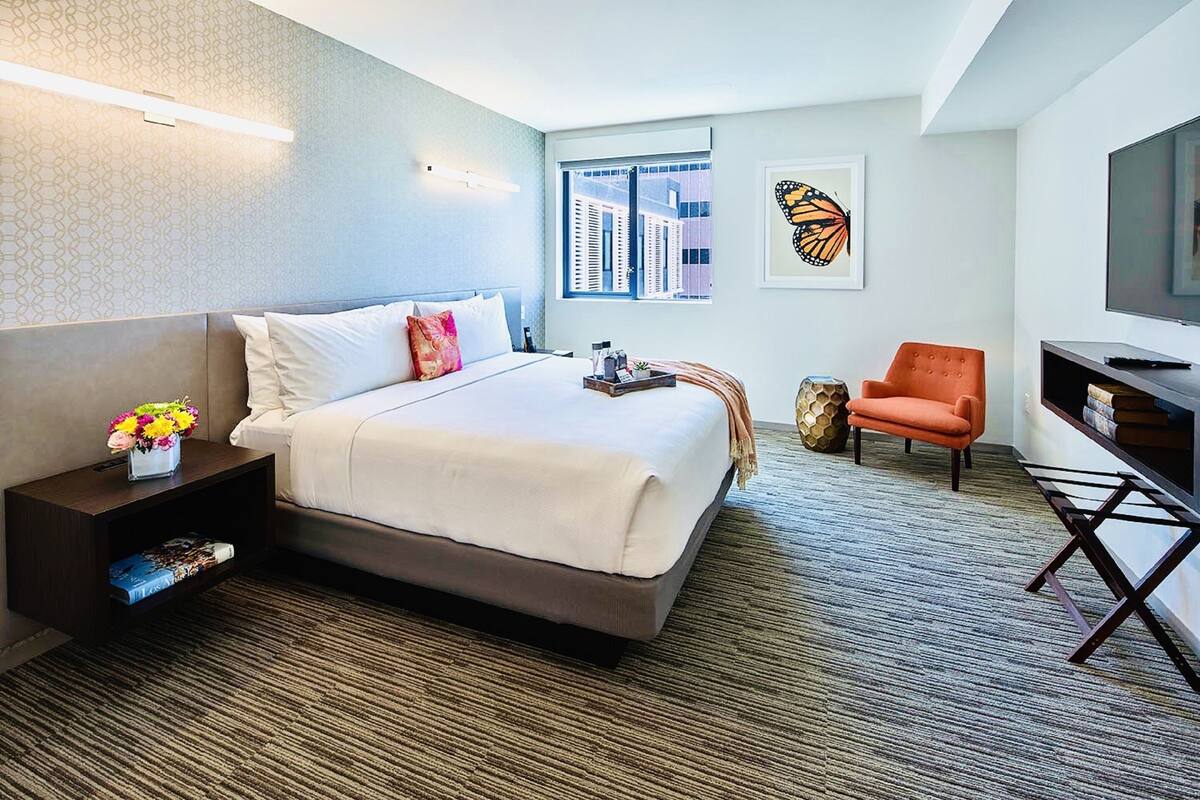 Une chambre d’hôtel avec un grand lit, une table de chevet ornée d’un vase de fleurs, une chaise, un téléviseur et une fenêtre donnant sur la ville.