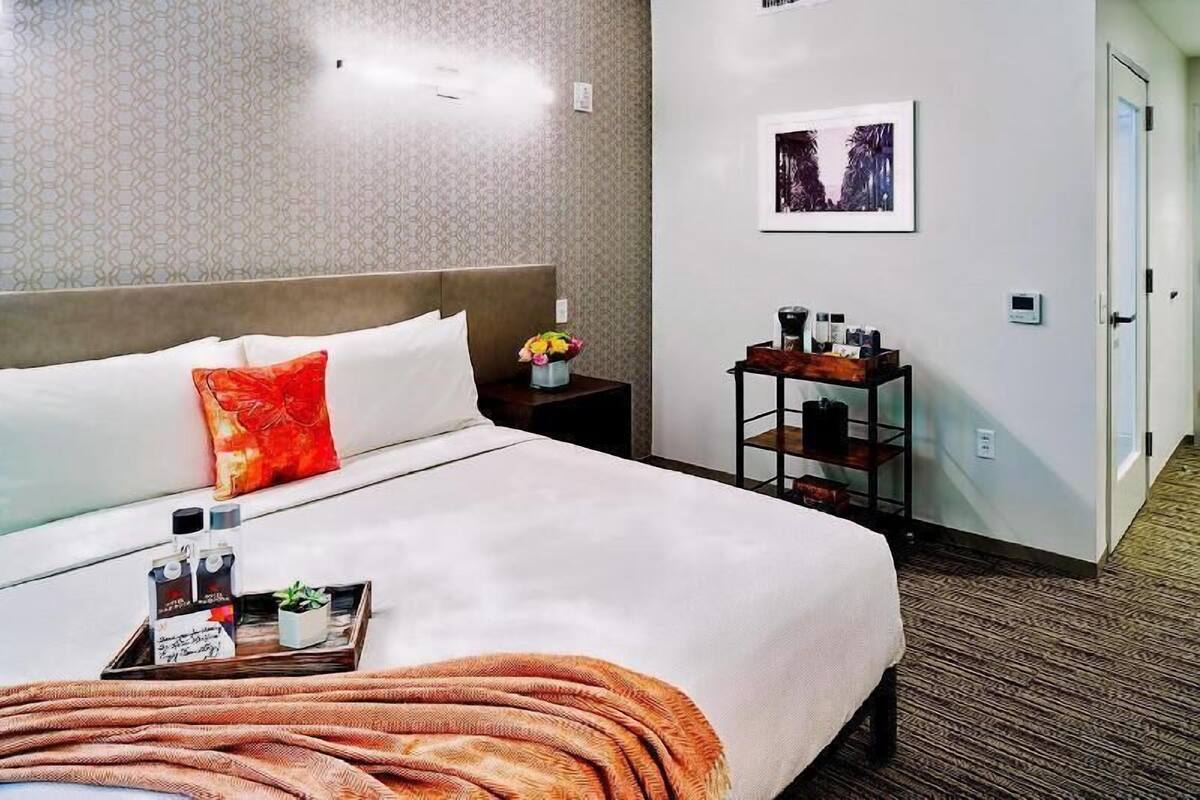 Une chambre d’hôtel avec un lit, une table de chevet ornée d’un vase de fleurs, un tableau encadré au mur et une petite table avec des articles de toilette.