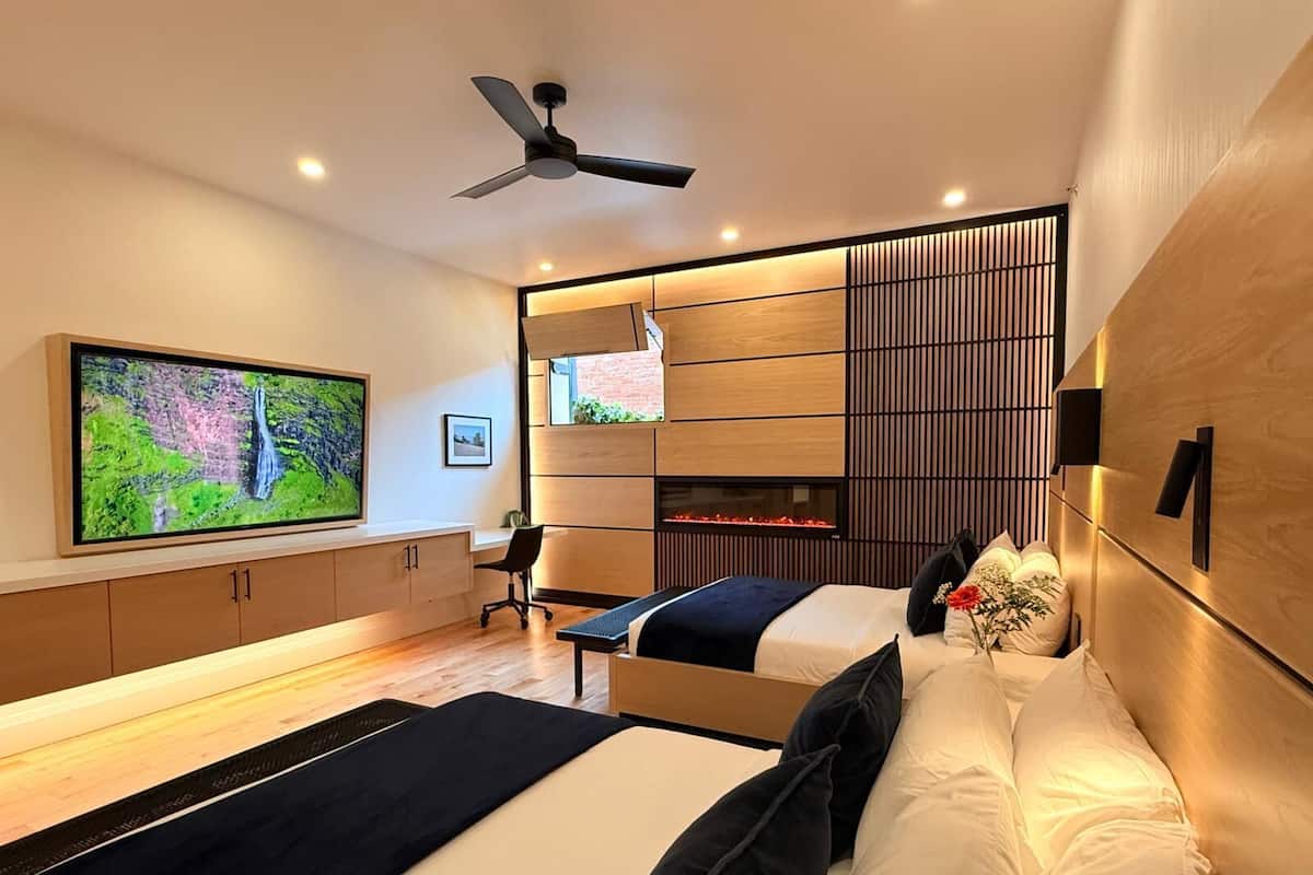 A modern bedroom with a large TV, a ceiling fan, a fireplace, and a bed with pillows.