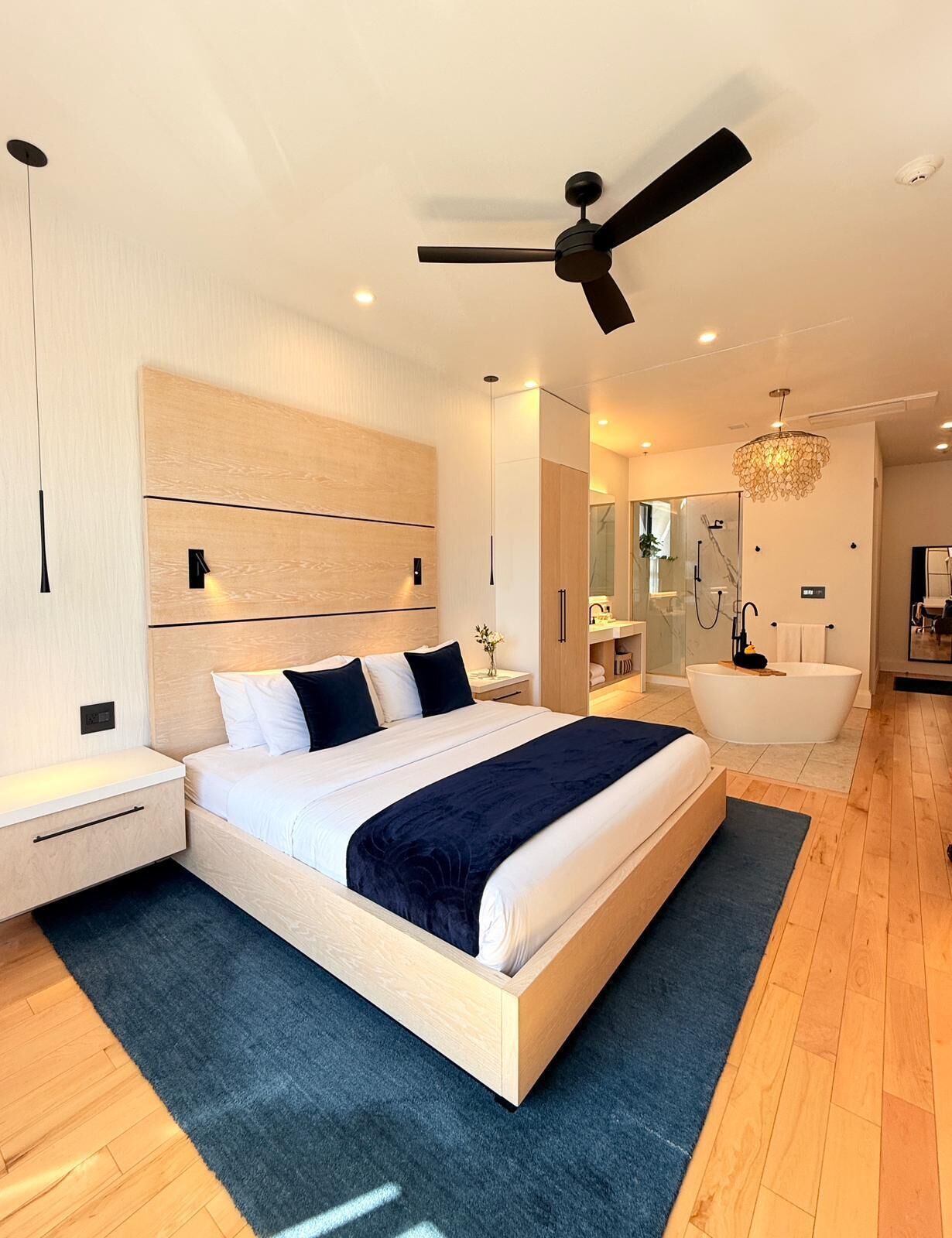 Une chambre moderne avec un grand lit, des tables de chevet, un ventilateur de plafond, un lustre et une salle de bain avec une baignoire.