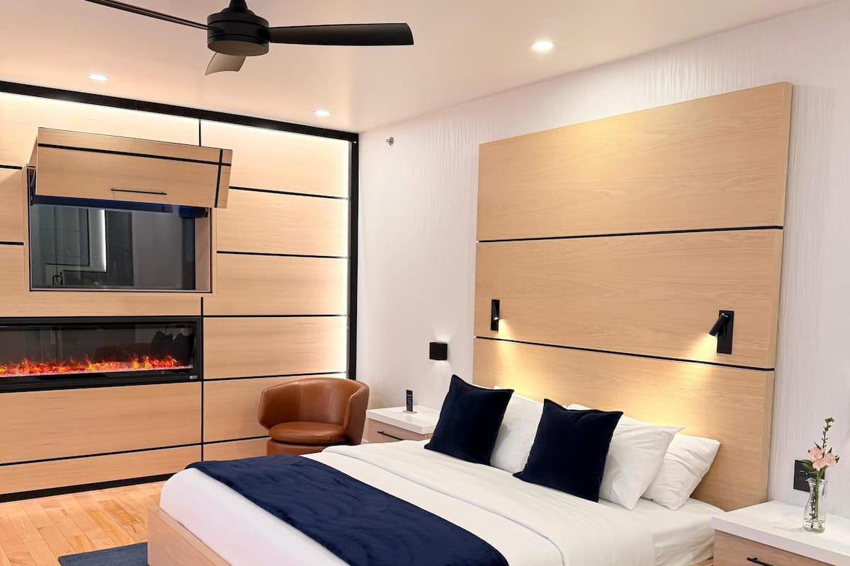 A modern bedroom with a bed, a ceiling fan, a fireplace, a chair, and a nightstand.