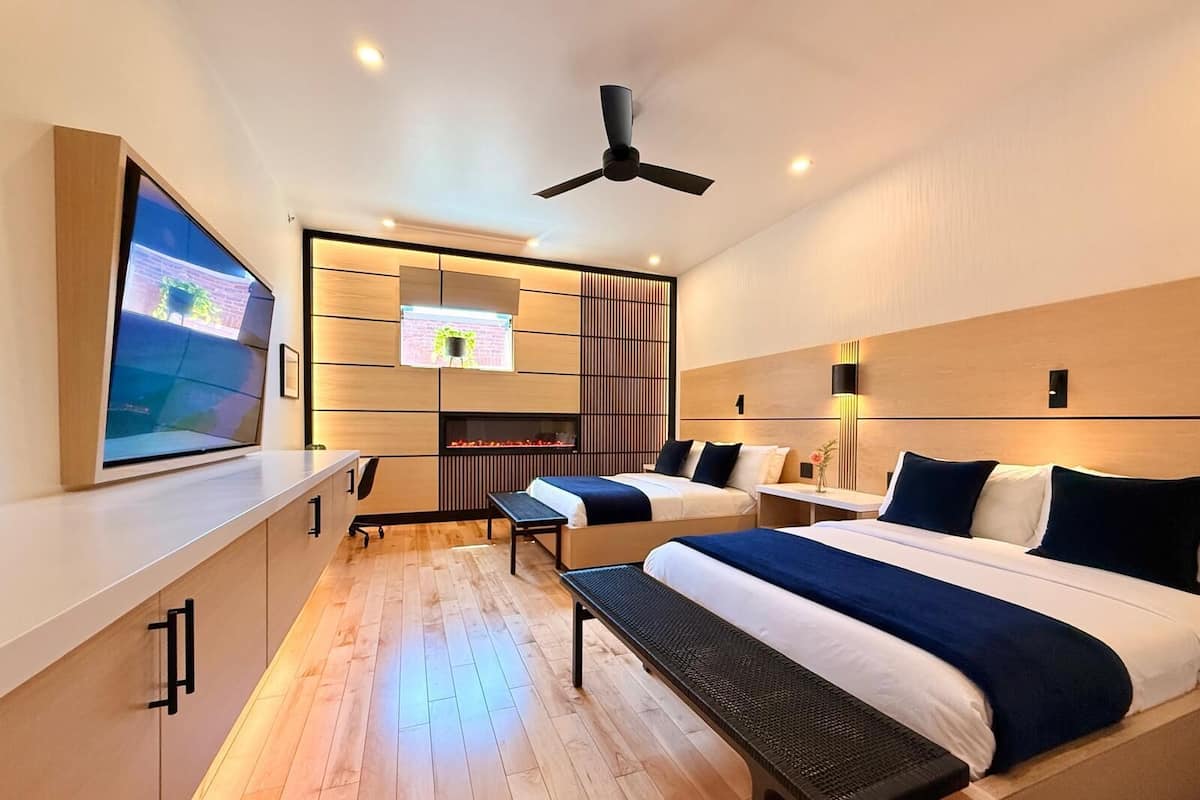 A modern hotel room with two beds, a desk, a TV, a ceiling fan, and a fireplace.