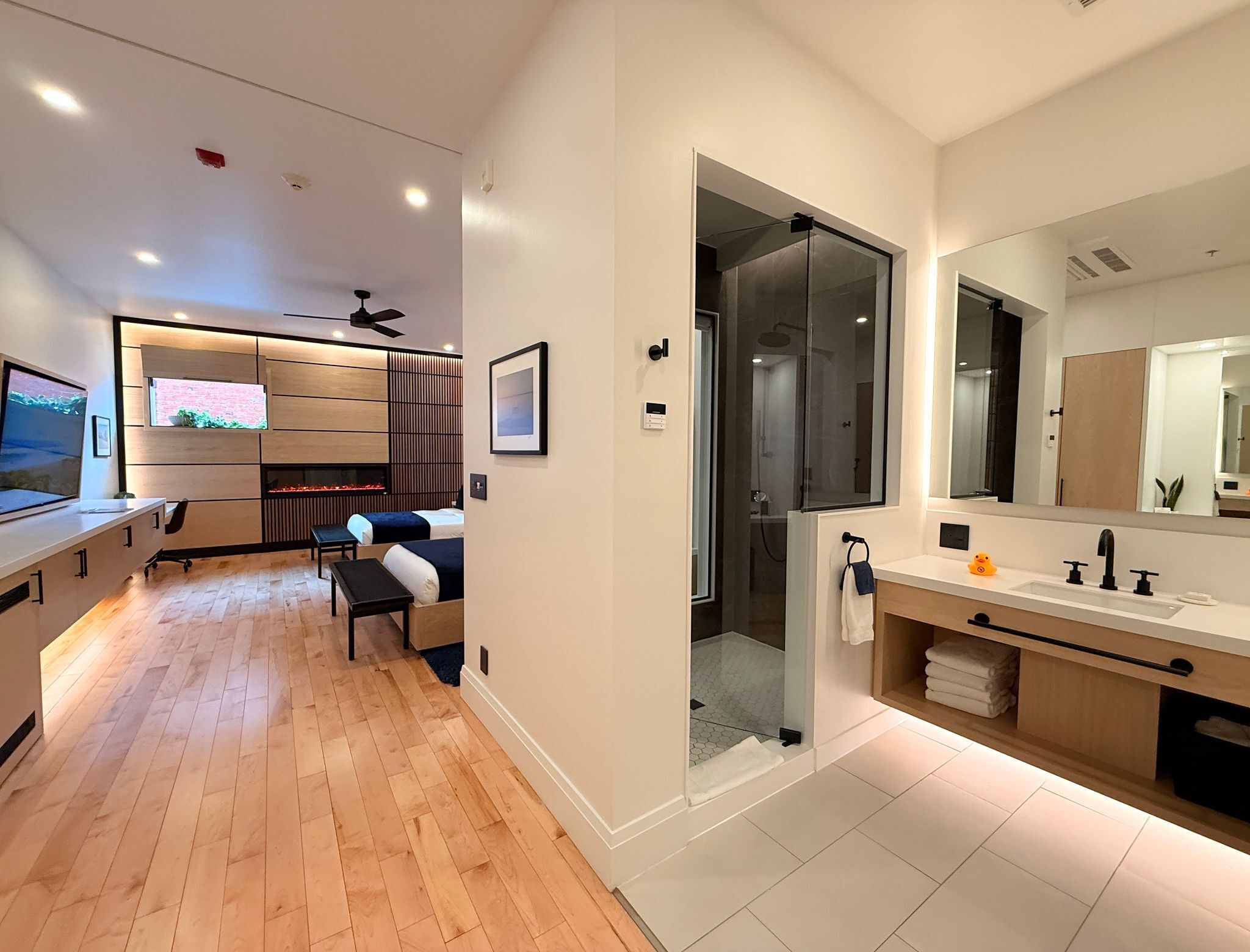 Ein modernes Hotelzimmer mit Holzboden, Fernseher, Bett, Schreibtisch, Dusche und Badezimmer mit großem Spiegel.