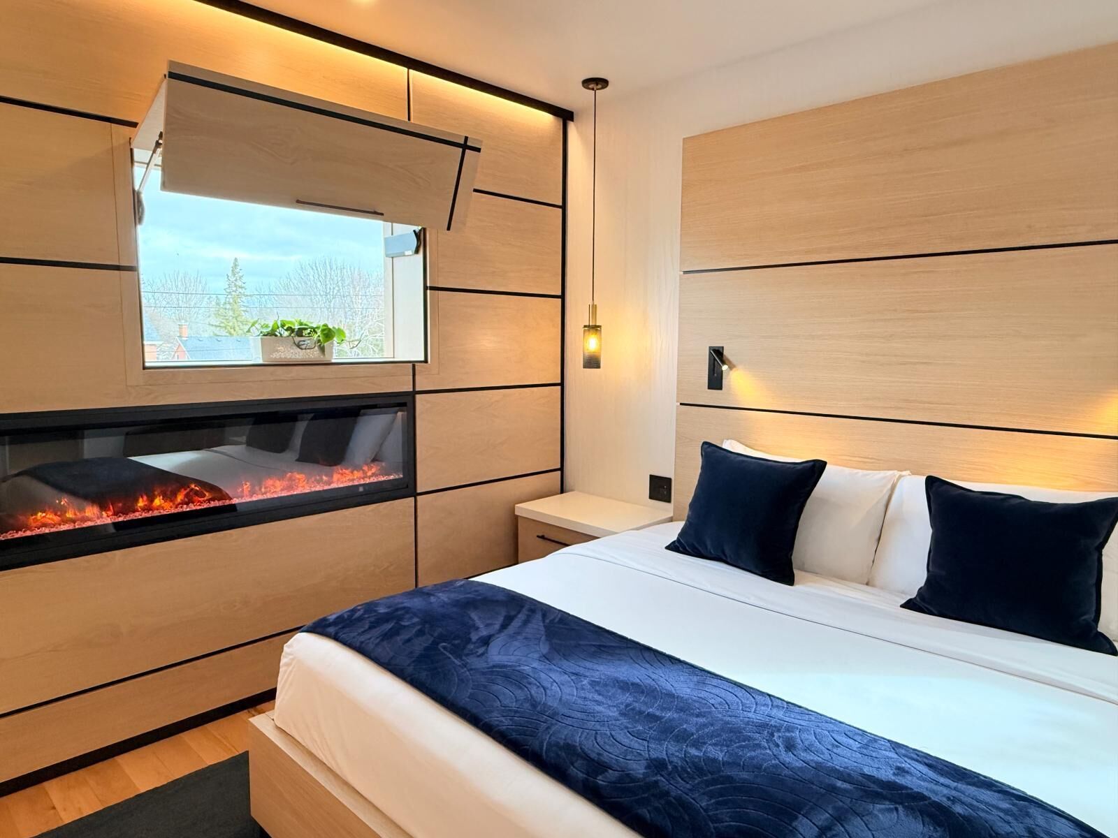 Une chambre à coucher moderne avec une cheminée intégrée, un grand lit avec une literie bleu foncé, et une vue sur un jardin, visible à travers une fenêtre située au-dessus de la cheminée.