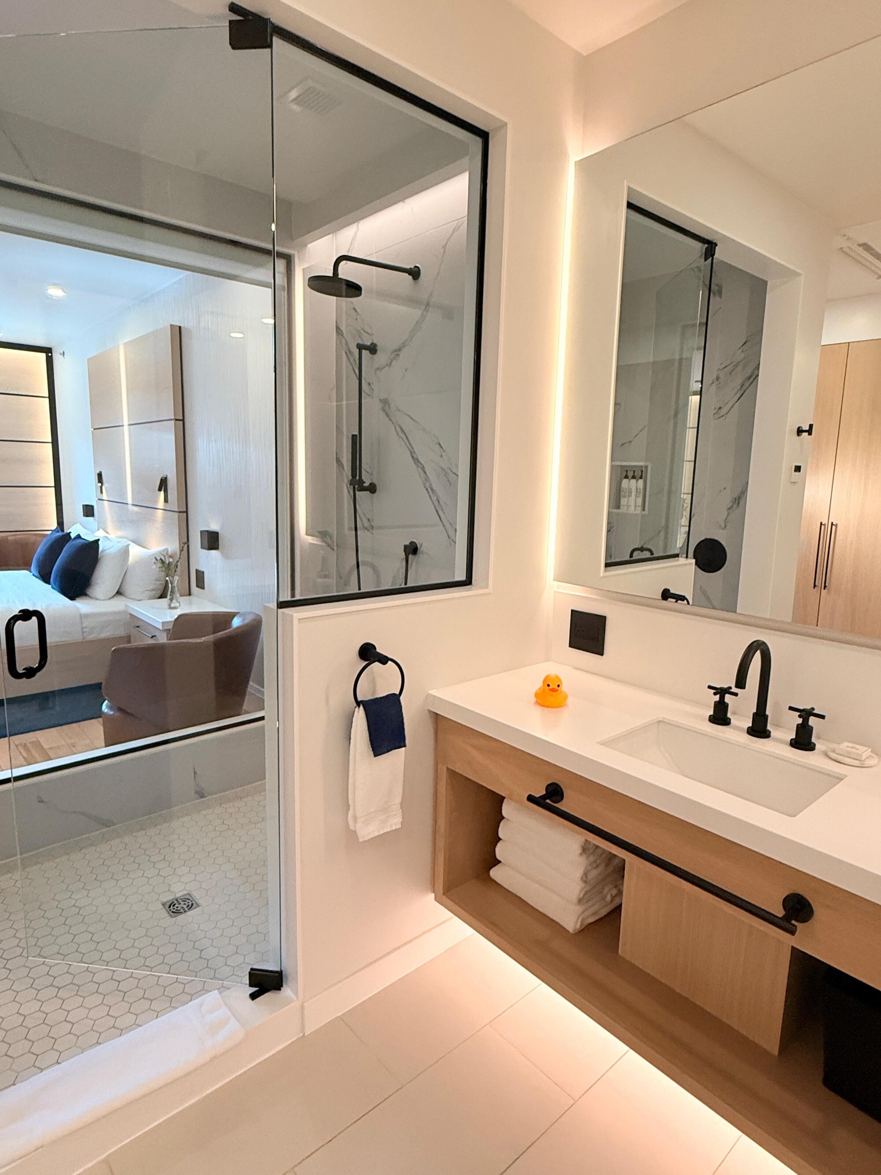 A modern bathroom with a glass shower enclosure, a large mirror, and a wooden vanity with a sink and towel rack.
