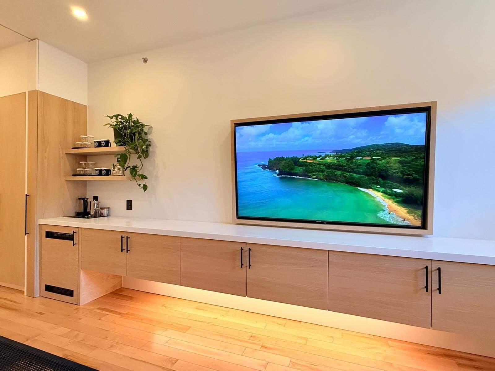 A modern kitchen with a large flat-screen TV mounted on the wall, wooden cabinets, and a countertop with a coffee maker and jars.