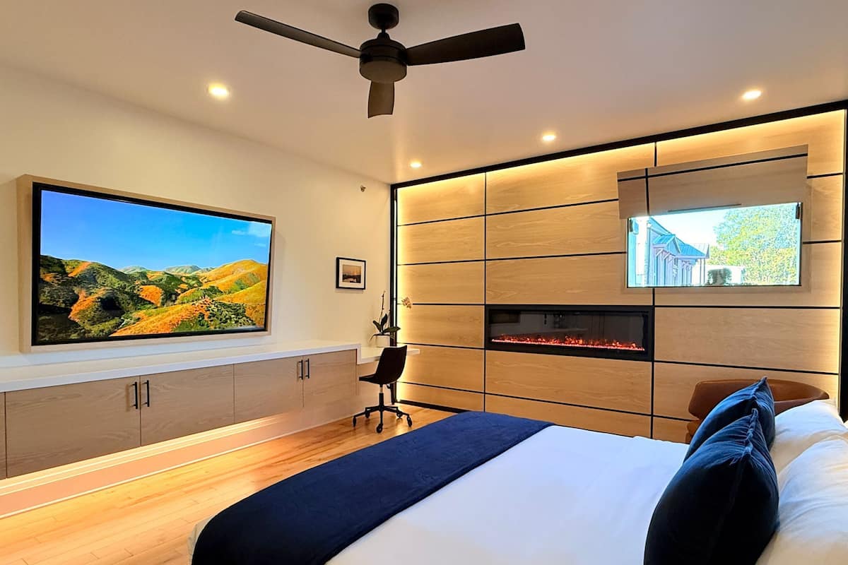 A modern bedroom with a large bed, a TV mounted on the wall, a ceiling fan, and a fireplace.