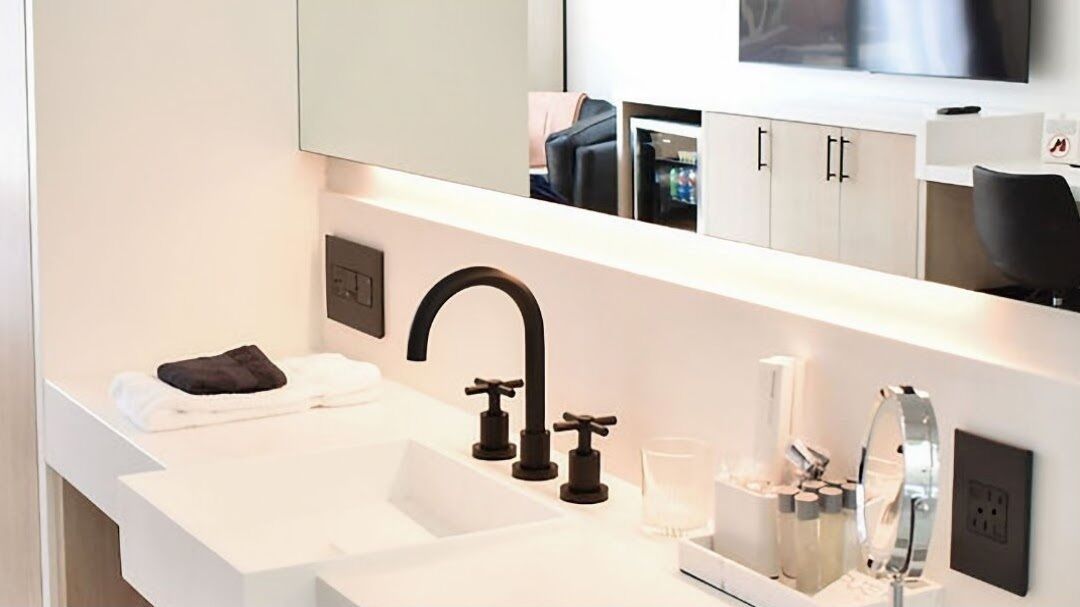 A modern bathroom with a white sink, black faucets, a large mirror, and a television above the mirror.