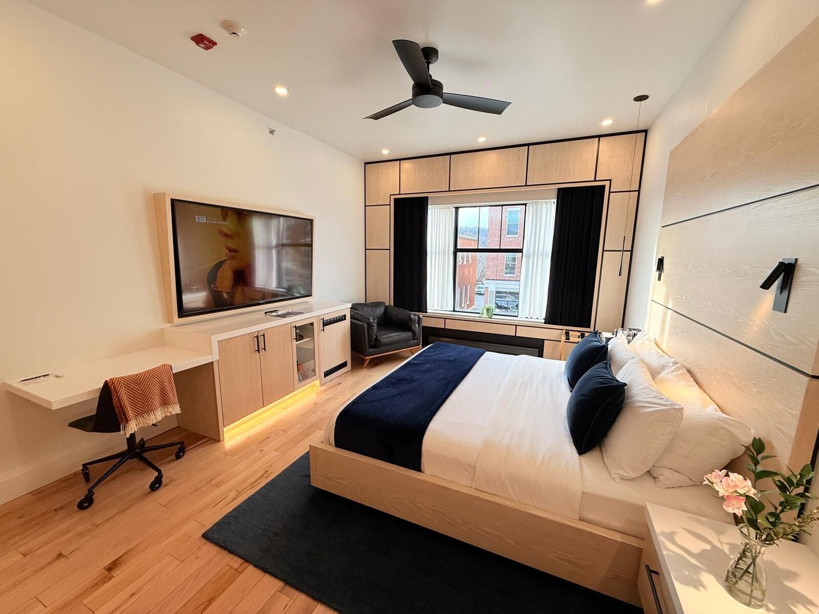 A modern hotel room with a large bed, a desk with a chair, a flat-screen TV, and a ceiling fan.