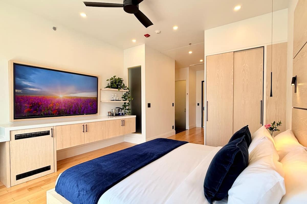 A modern bedroom with a large bed, a flat-screen TV mounted on the wall, wooden cabinets, and a ceiling fan.