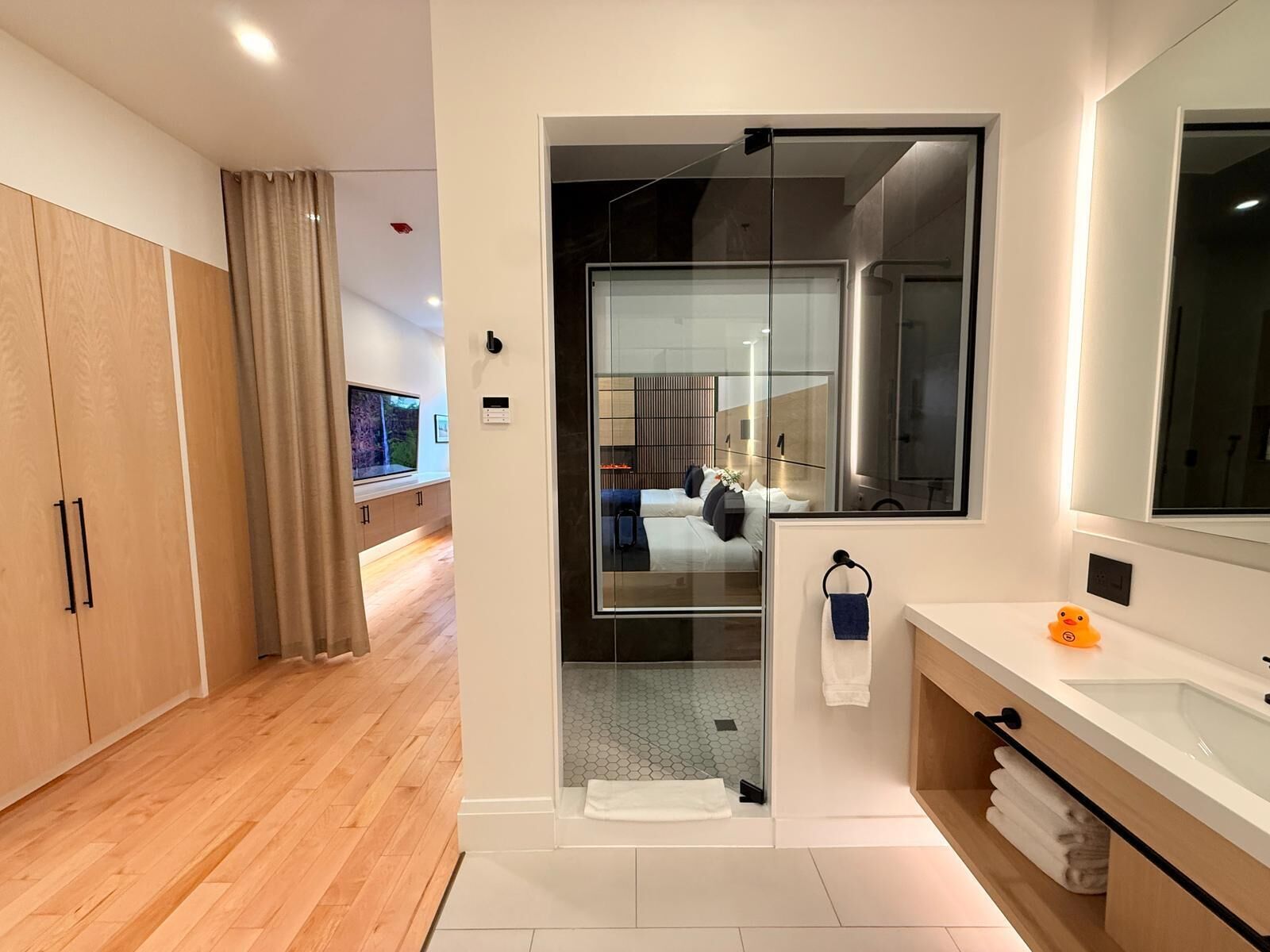 A modern hotel room with a wooden wardrobe, a large mirror, a glass shower enclosure, and a bathroom with a sink and towel rack.