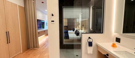 A modern hotel room with a wooden wardrobe, a large mirror, a glass shower enclosure, and a bathroom with a sink and towel rack.