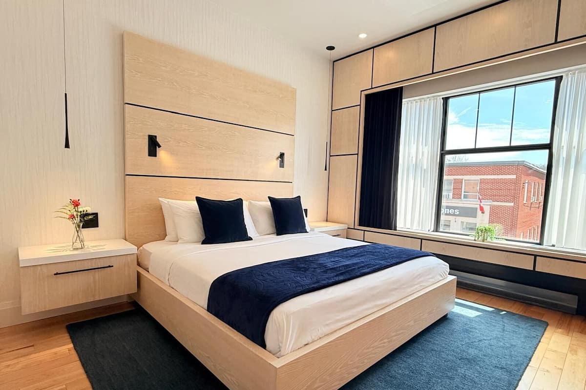 A modern bedroom with a large bed, wooden headboard, bedside tables, a window with a view of buildings, and a small plant on the windowsill.