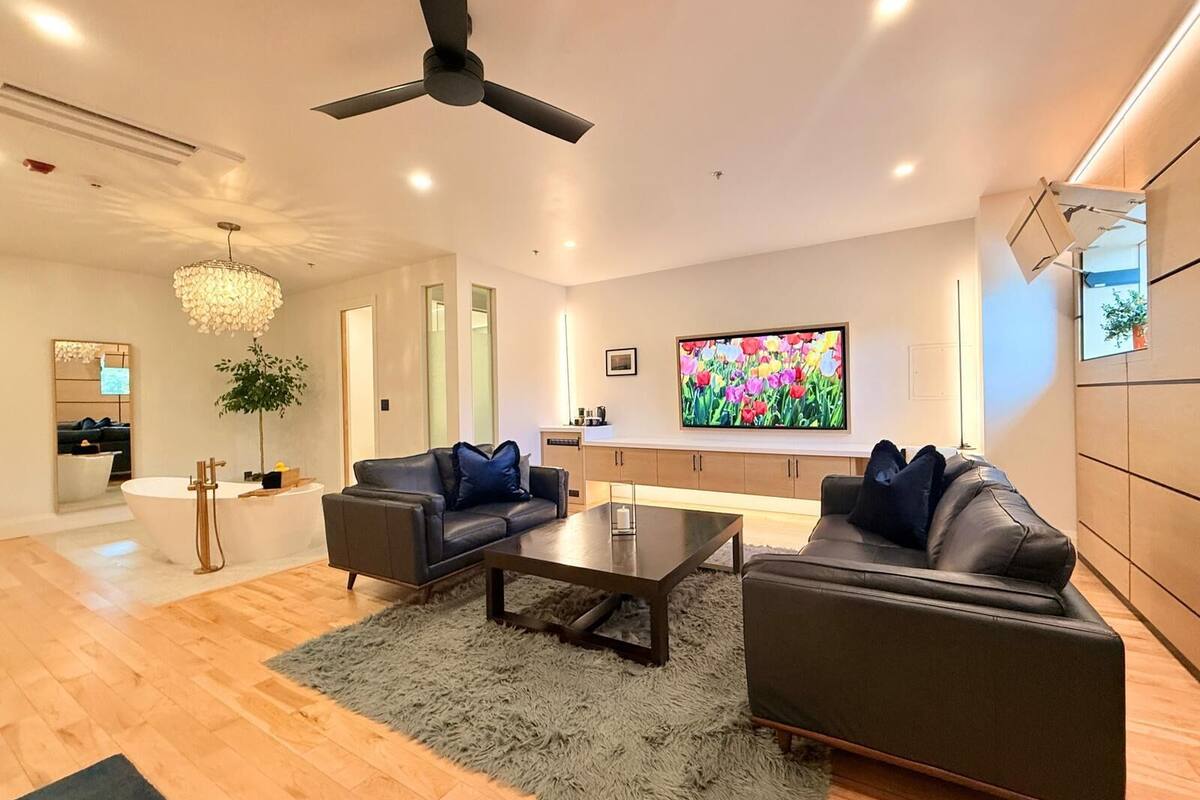 A modern living room with a large flat-screen TV, a ceiling fan, and a chandelier.