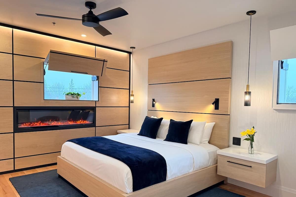 A modern bedroom with a bed, a TV mounted on the wall, a fireplace, and a ceiling fan.