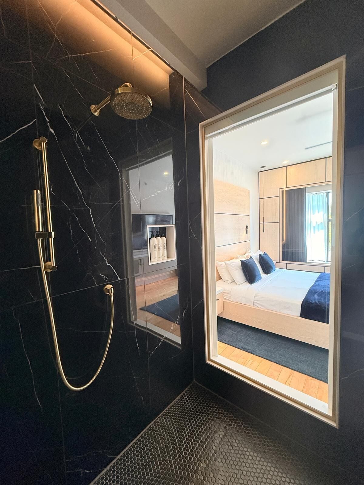 Un baño moderno con un espejo grande, una ducha con cabezal dorado y vistas a un dormitorio con cama y ventana.