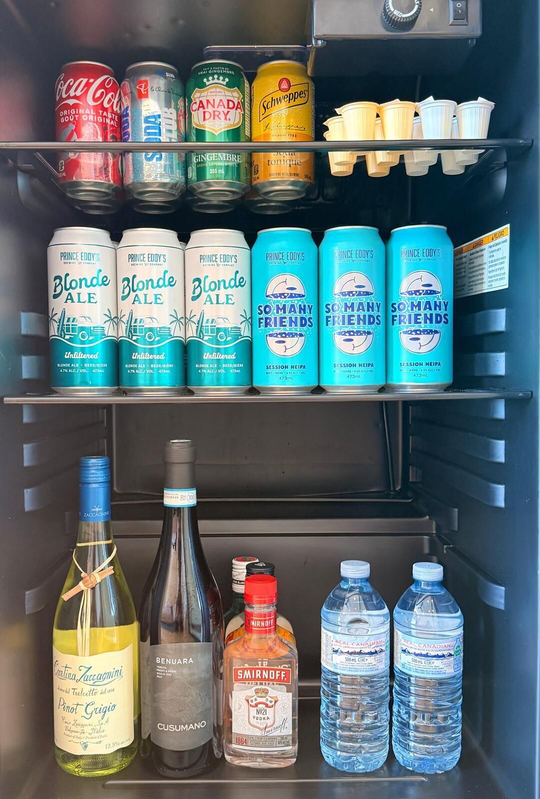 A refrigerator stocked with beverages including cans of Coca-Cola, Canada Dry, Schweppes, and Prince Eddy's Blonde Ale.