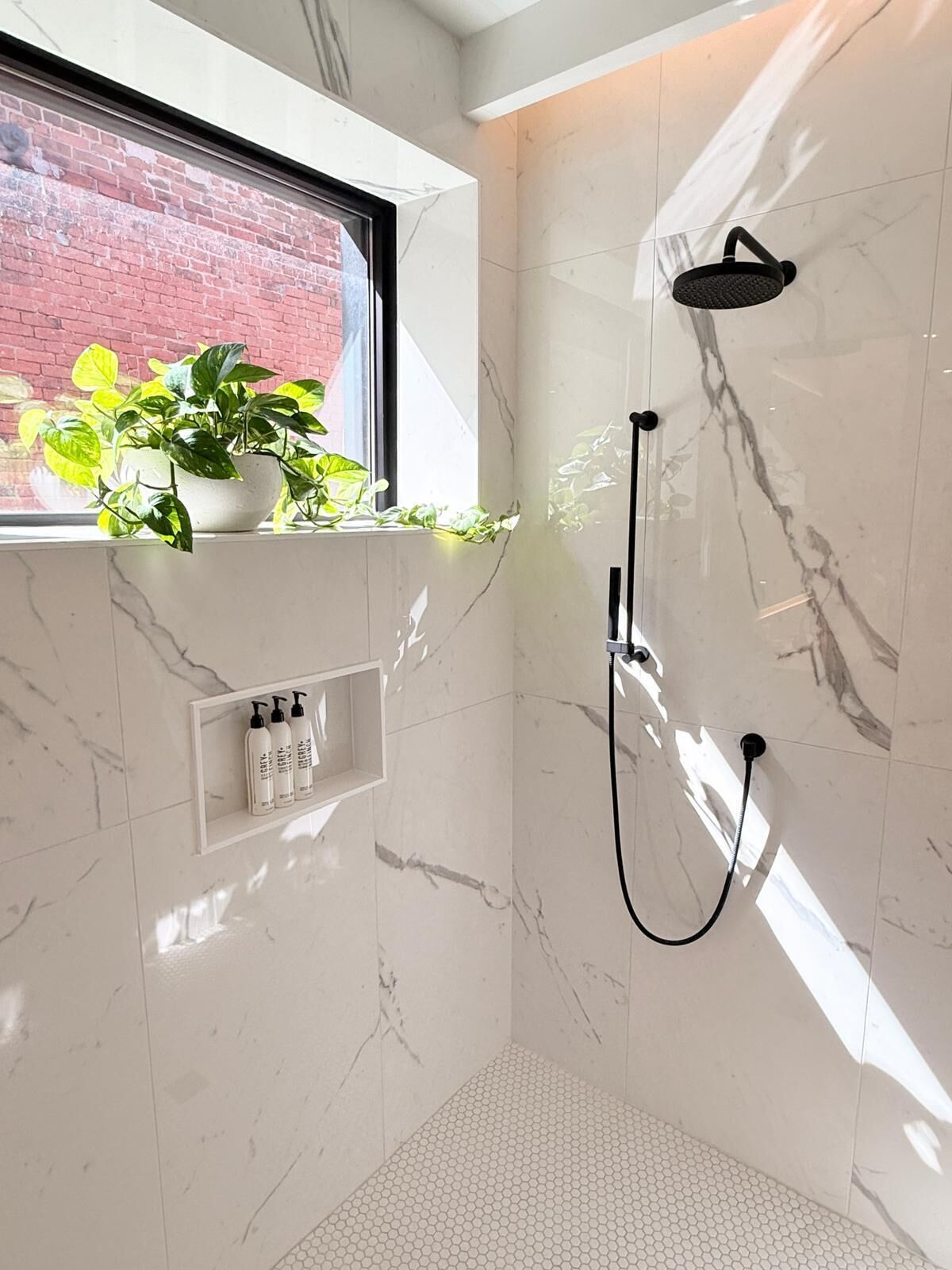 A modern bathroom with a shower, a window with a view of a brick wall, and a shelf with toiletries.