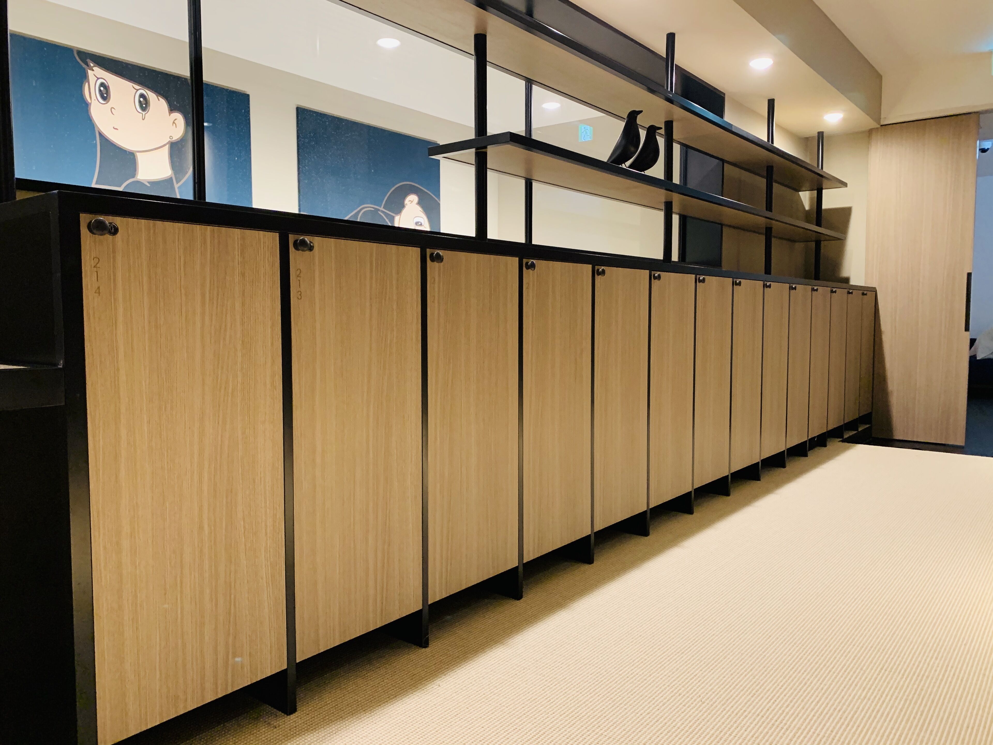 A row of wooden lockers with metal handles, a black metal shelf above, and a blue wall with cartoon characters.