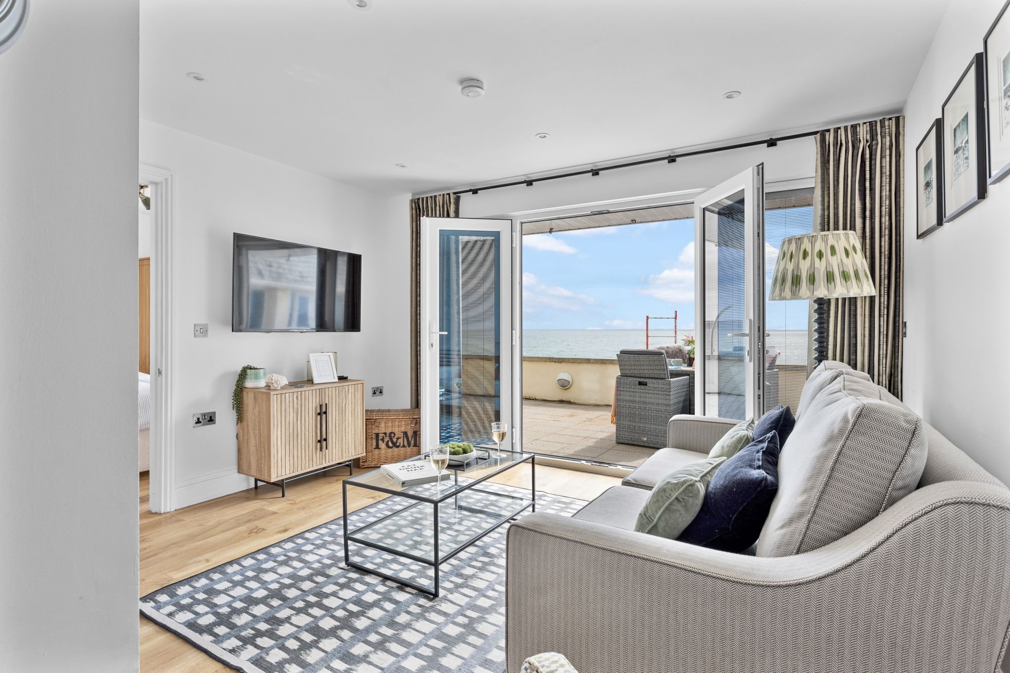 Un salon moderne avec un canapé, une table basse et un téléviseur fixé au mur. On peut également apercevoir un meuble en bois et un fauteuil en osier.