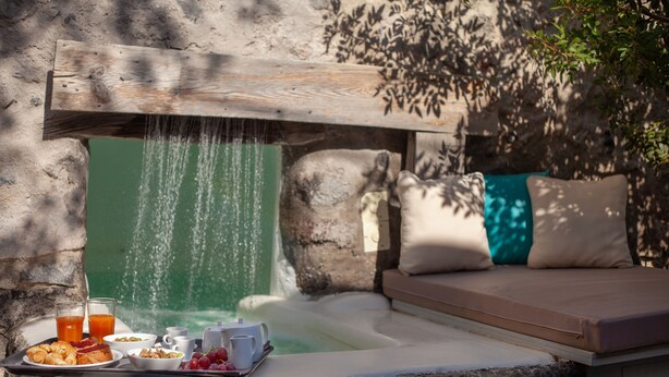 A hot tub with a wooden bench and cushions, a tray with breakfast items, and a waterfall feature.