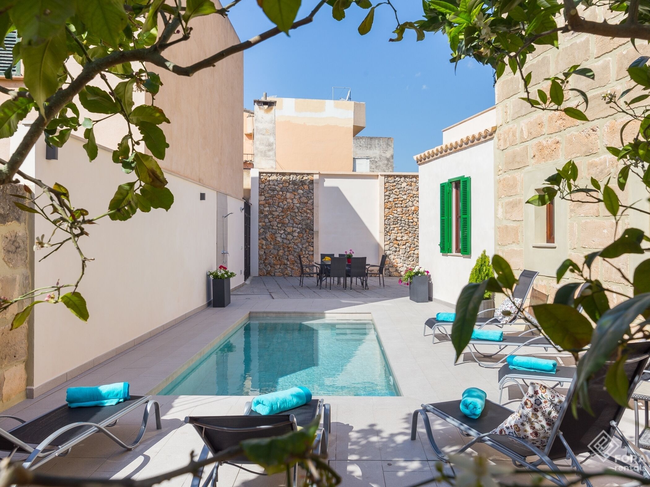 A poolside area with lounge chairs and a table, surrounded by stone walls and a clear blue sky.
