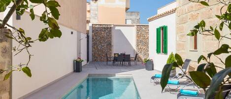 A poolside area with lounge chairs and a table, surrounded by stone walls and a clear blue sky.