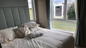 A neatly made bed with a headboard, pillows, and a window with curtains. - Brand new luxury holiday home in one of the best holiday parks in the country. (Perranporth)