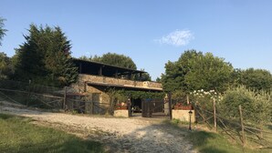 A rustic stone building with a wooden balcony, surrounded by greenery and a gravel path leading to a gated entrance. - Colline Toscane, beautiful two-room apartment for 2/4 people, lovely view and swimming pool (Casole d’elsa, siena)