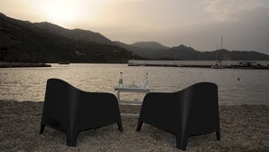 Dos sillas negras orientadas hacia una mesa con botellas, con vista a una masa de agua tranquila y montañas al fondo.