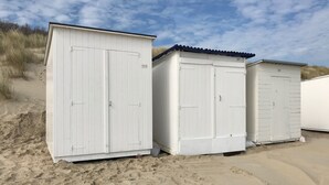 Tres cobertizos blancos con puertas metálicas, numerados 303 y 305, en una playa arenosa con vegetación escasa.