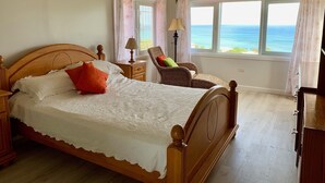 Een slaapkamer met een houten bed, een rieten stoel, een nachtkastje, een lamp en uitzicht op de oceaan door grote ramen.