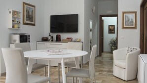 Un salon moderne avec une table à manger blanche, quatre chaises, un téléviseur à écran plat et un coin cuisine équipé d’un four à micro-ondes et d’un grille-pain.