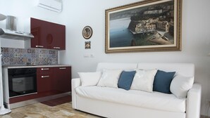 Une cuisine moderne équipée d’une cuisinière, d’un four et de meubles de rangement rouges. Un canapé blanc avec des coussins bleus est placé contre le mur, et un tableau encadré représentant une ville côtière est accroché au-dessus.
