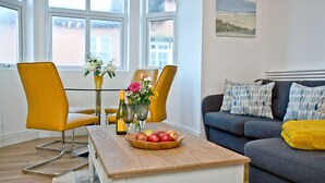 A modern living room with a dining area, featuring a wooden coffee table with fruit, yellow chairs, and a grey sofa with patterned cushions. - 5 At The Beach - Beachfront, Parking, Pets welcome (Torcross)