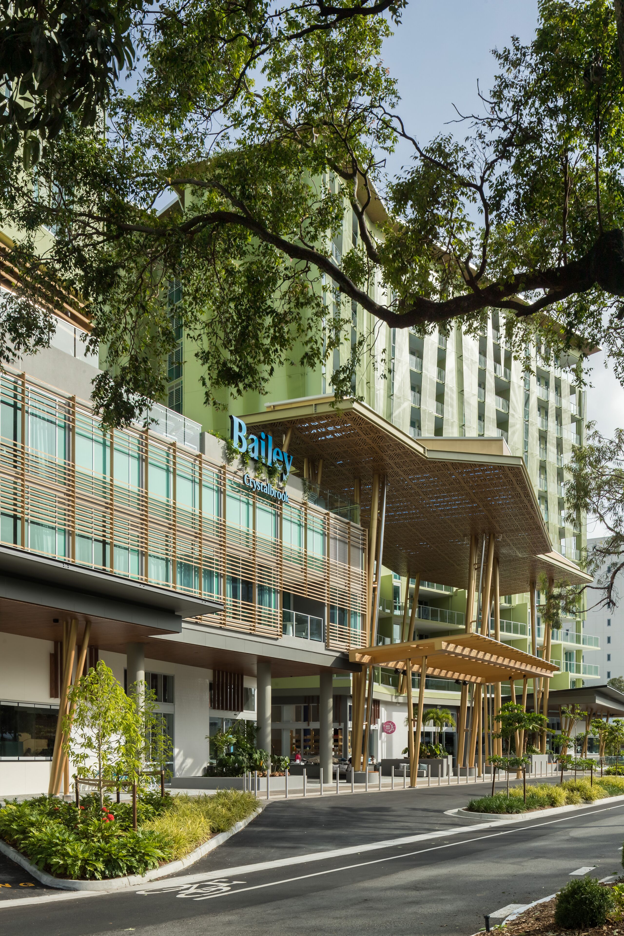 A modern hotel with a prominent "Bailey Crystalbrook" sign, featuring a unique wooden canopy structure and lush greenery around the entrance.
