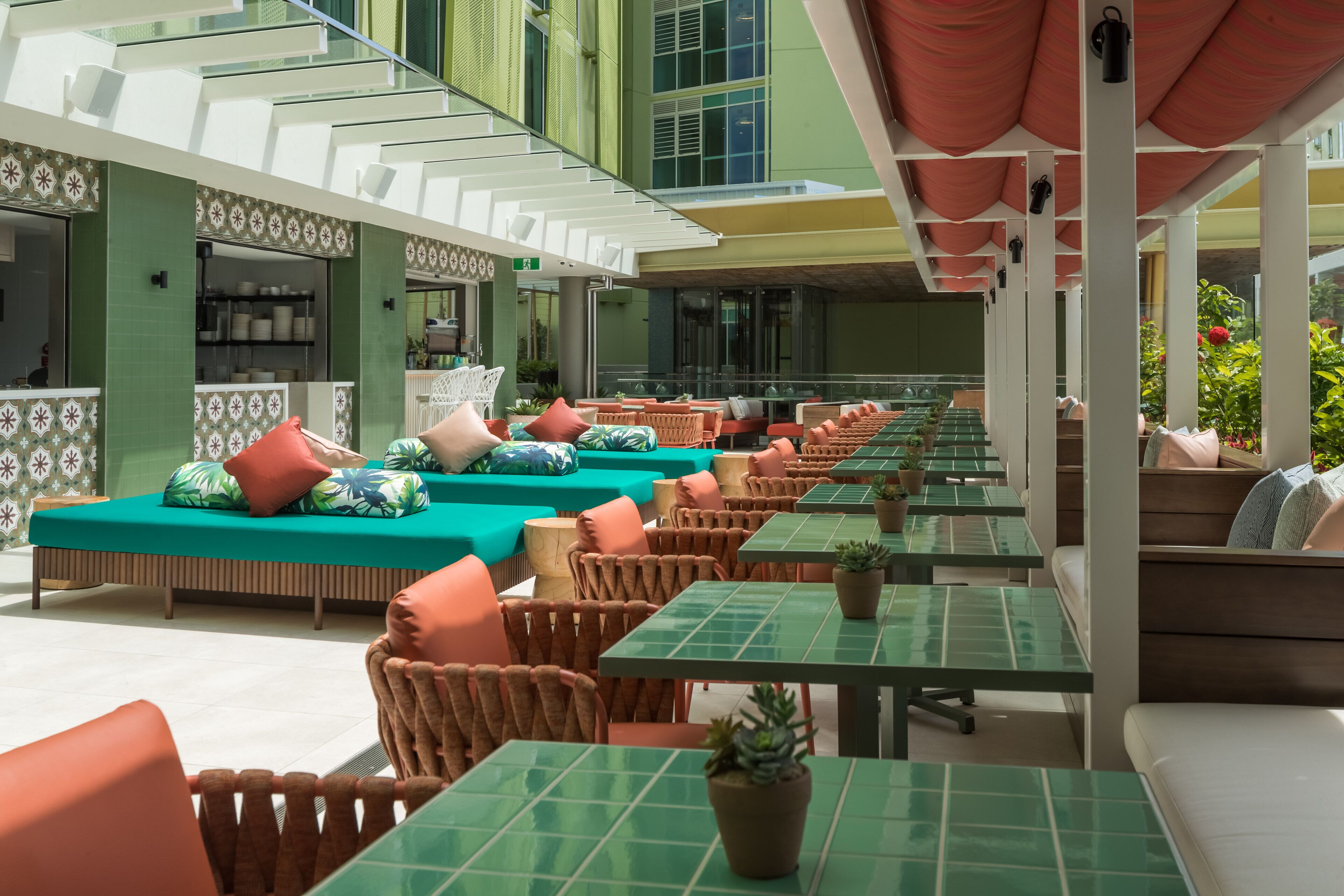 A covered outdoor seating area with green tables and chairs, a sofa with cushions, and a kitchen area in the background.