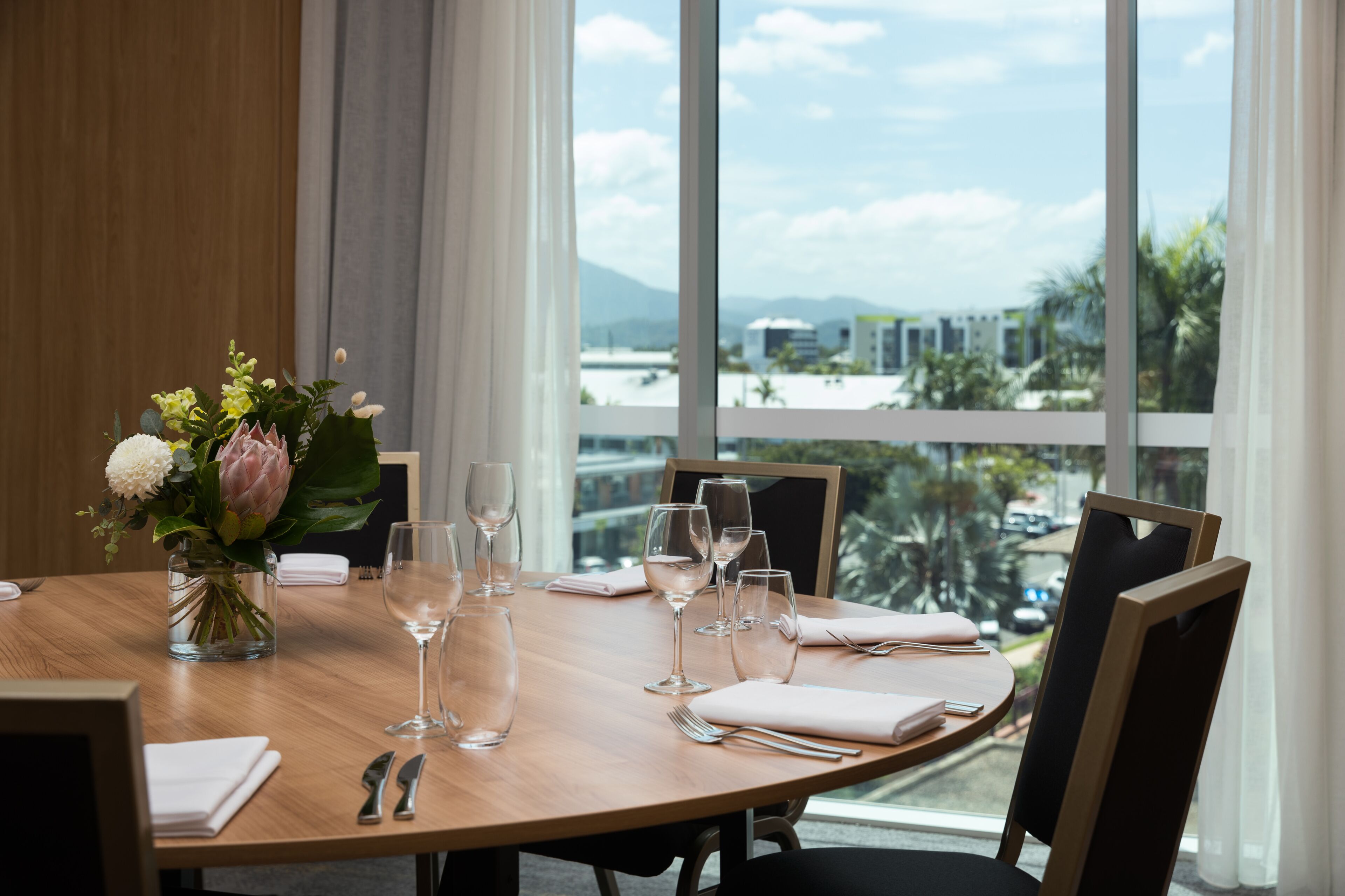 Une table à manger dressée pour six personnes, avec six verres à vin, un vase rempli de fleurs et une vue sur la ville par de grandes fenêtres.