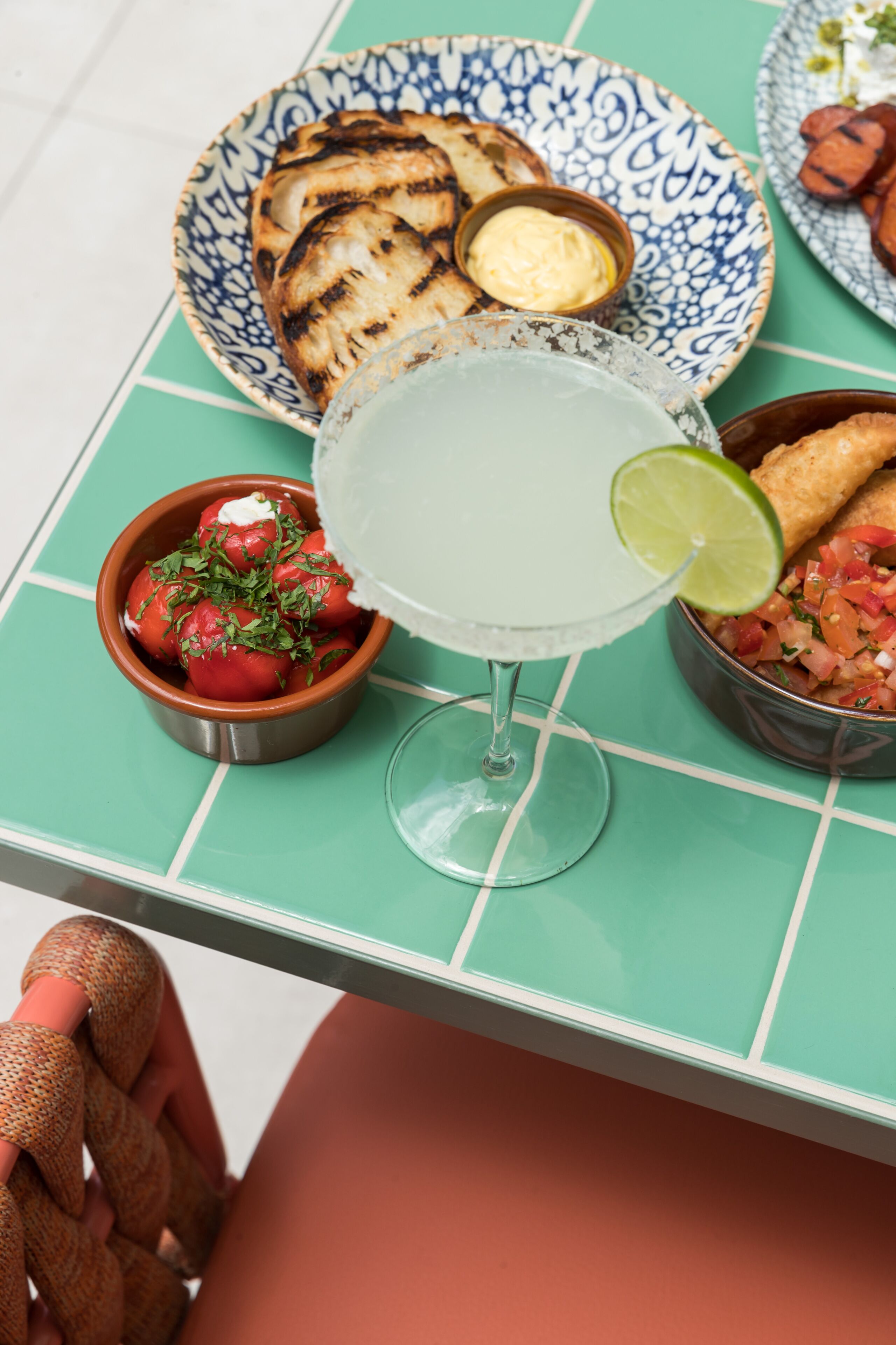 Una mesa con mantel verde, una copa de margarita con una rodaja de lima, un cuenco de salsa, pan a la parrilla y un bol de tomates con hierbas.
