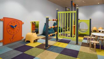A children's play area with a colorful checkered floor, a large orange clock, and a play structure with a green gate.