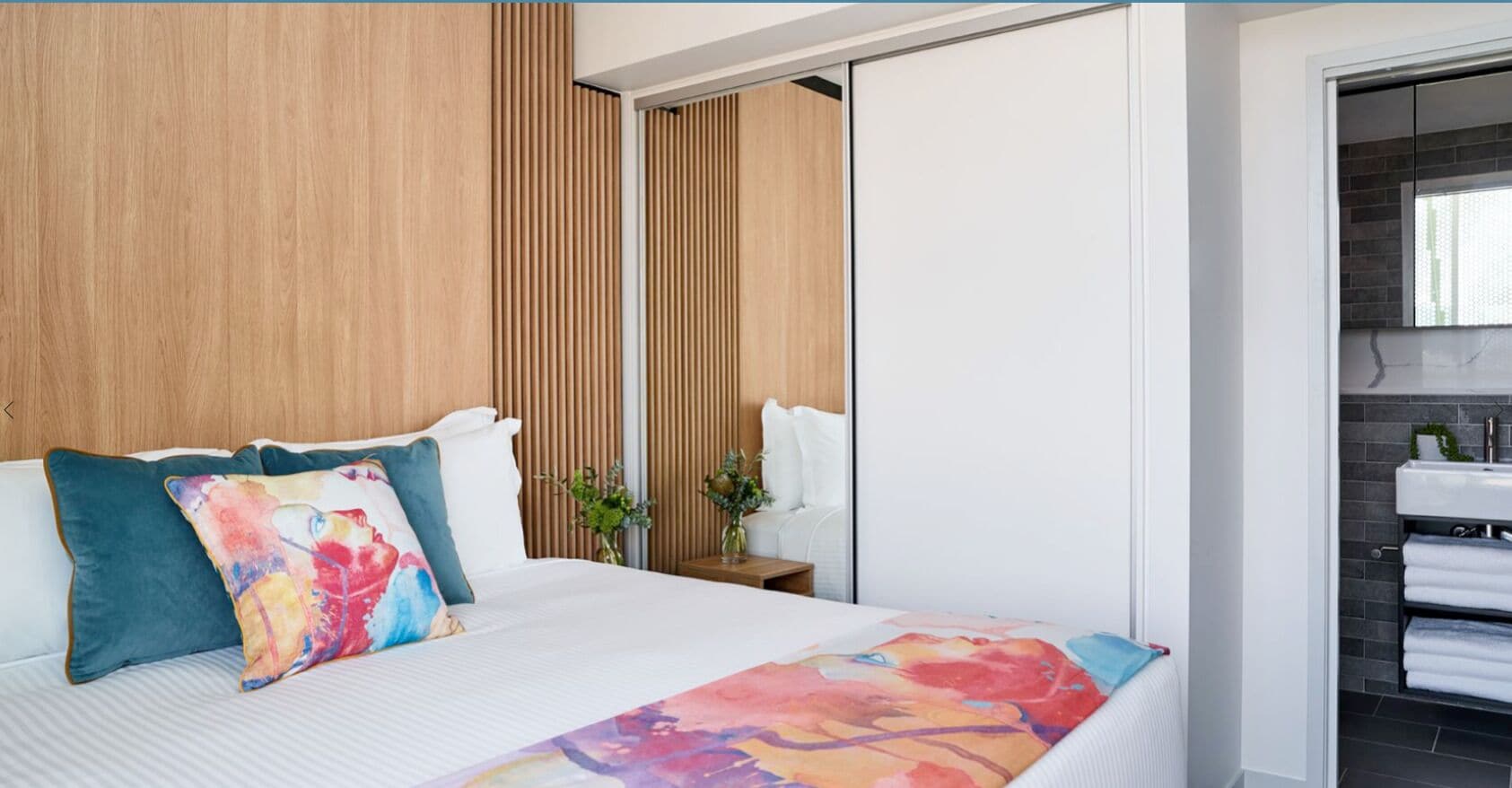 A hotel room with a bed, a wooden panel wall, a white bedside table with a vase, and a bathroom visible through an open door.