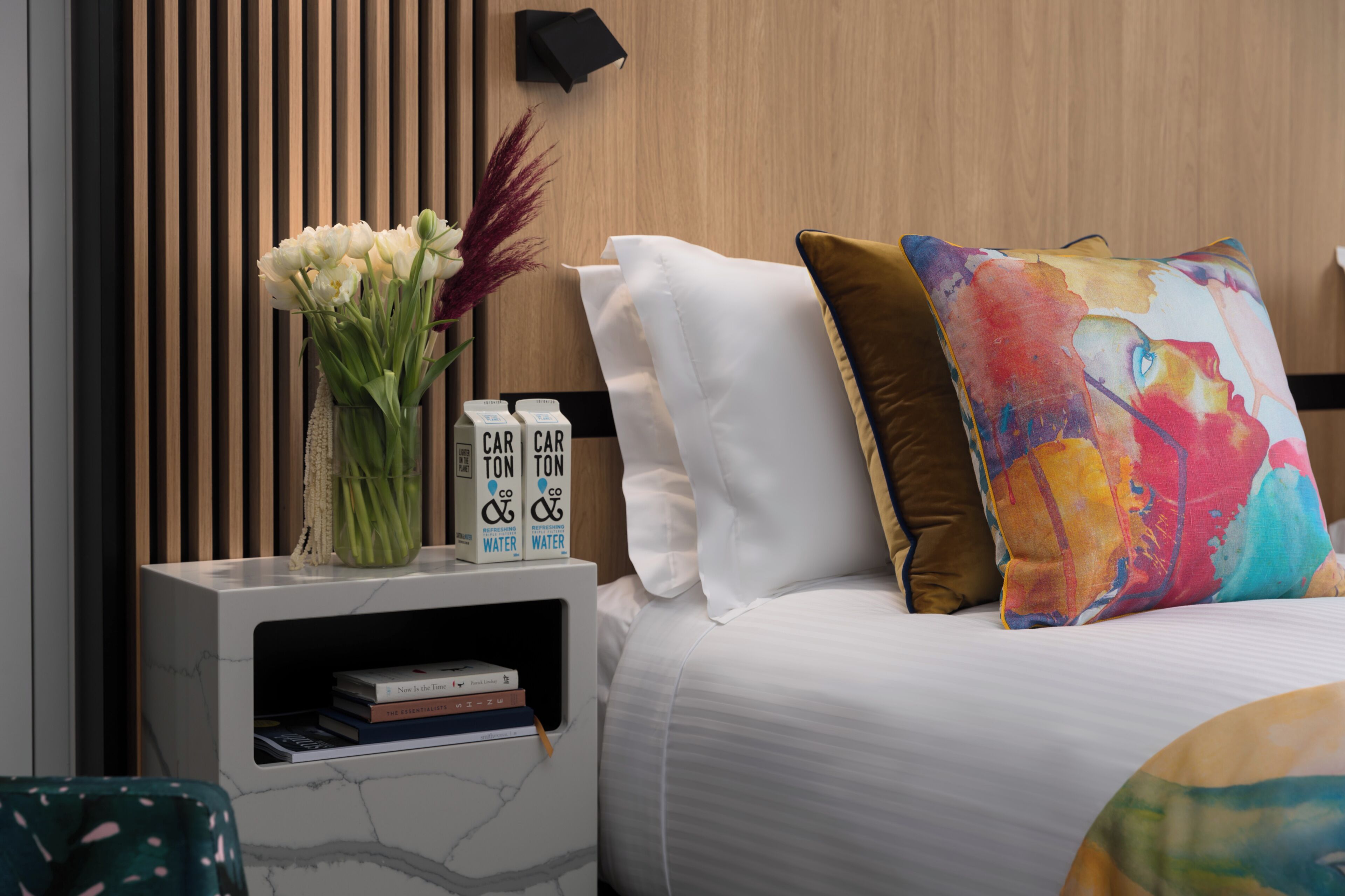 Un dormitorio ordenado con cama, almohadas, una mesita de noche con un jarrón de flores y una estantería con libros y botellas de agua.