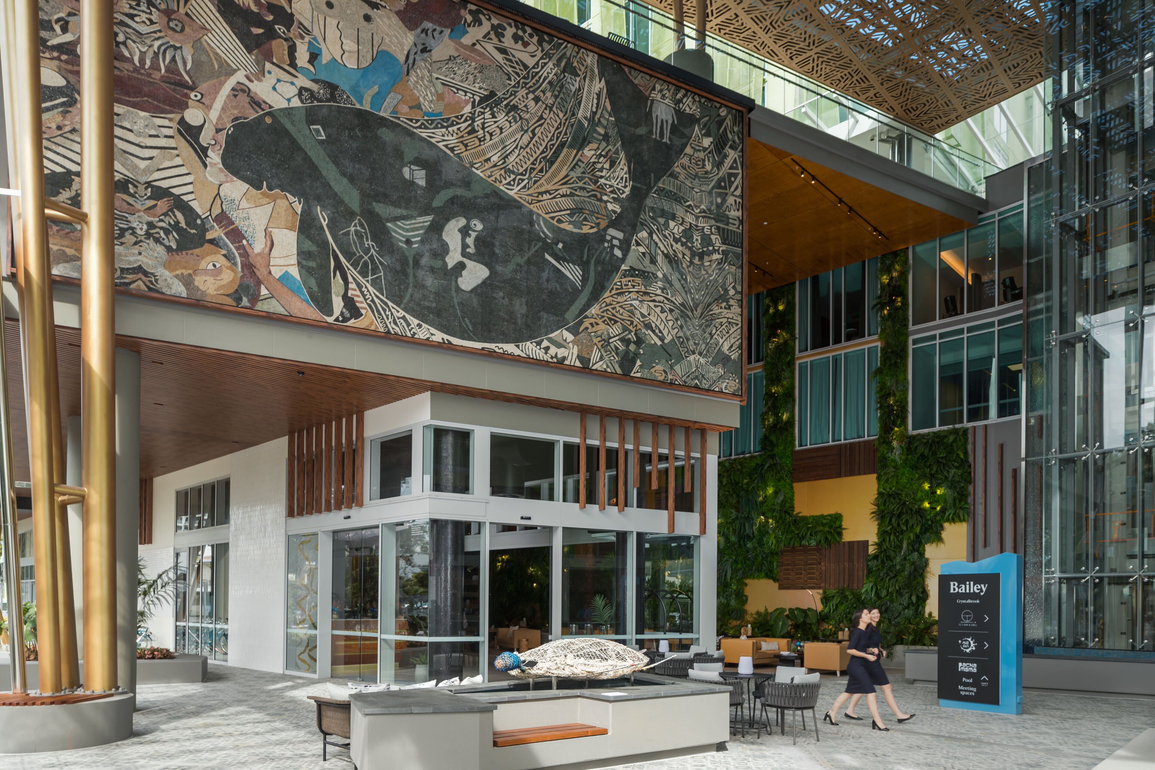 A modern hotel lobby with a large mural, glass walls, and a seating area.