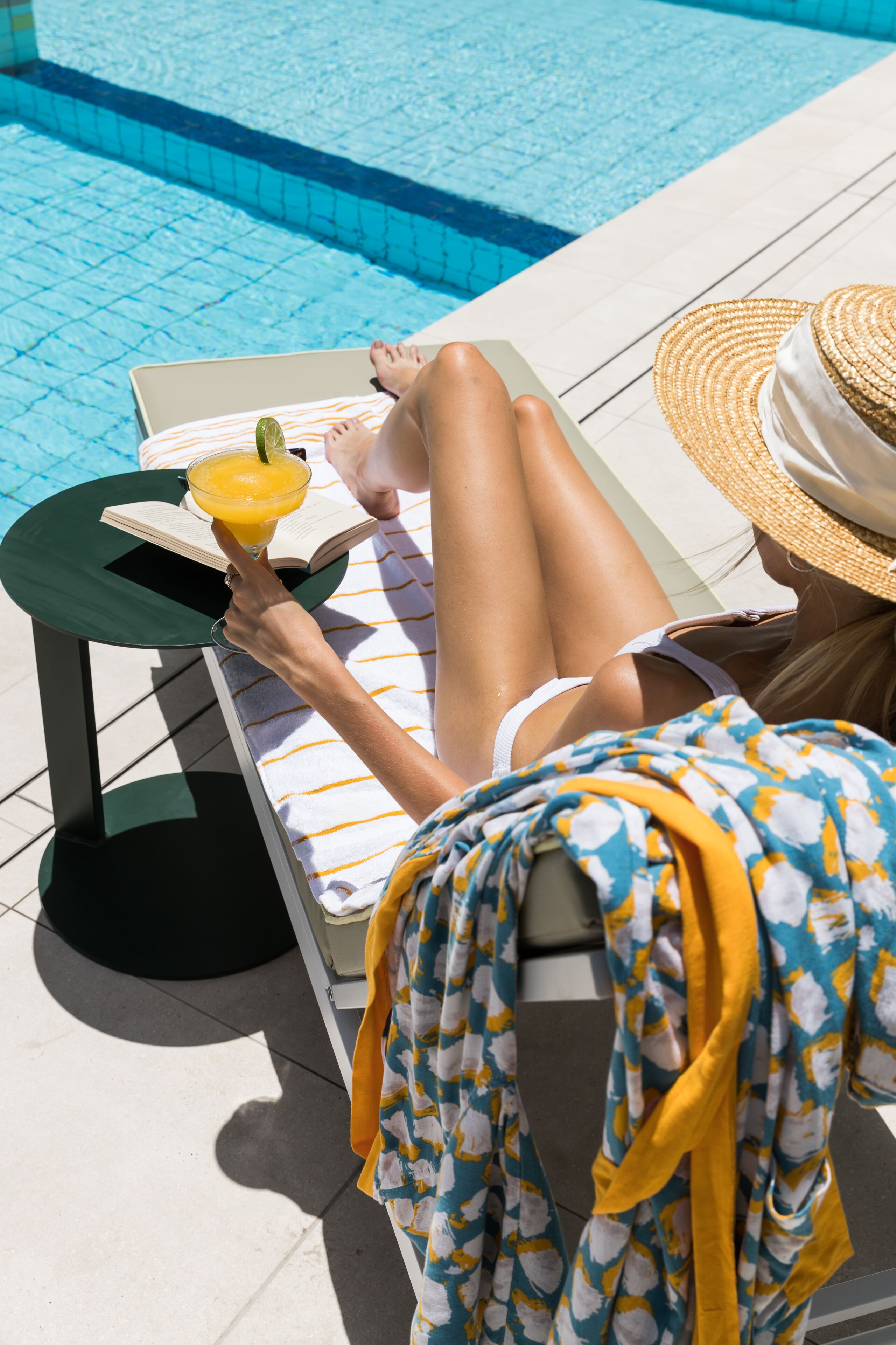 A person lounging by a poolside with a book and a drink.
